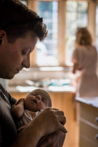 Dad hold newborn son while Mum is busy. Image by Sophie Ransome Lifestyle Photographer