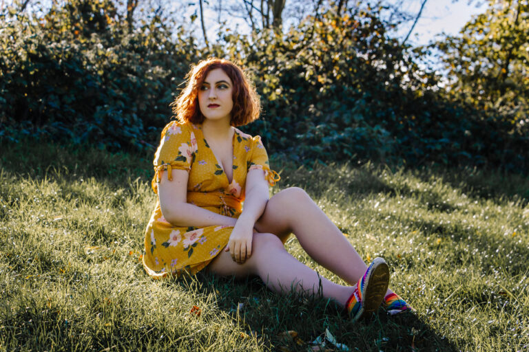 A teenager in a yellow dress sits on the grass in the sunshine during a portrait session with Sophie Ransome Lifestyle Photographer